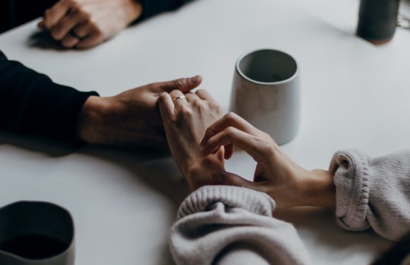 Two people hold hands across a white table, with mugs and a small potted plant nearby, near a window with natural light coming in.