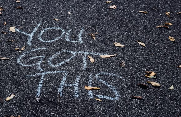 "Yellow chalk writing says 'You look great!' on a dark asphalt surface with scattered dry leaves."