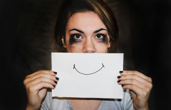 A young woman with smudged black makeup around her eyes holds a white paper with a simple smiley face drawing in front of her mouth, against a dark background.