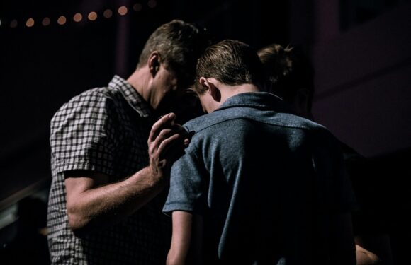 A group of people, including at least one man in a plaid shirt and a boy in a grey shirt, are gathered closely together indoors with dim lighting.