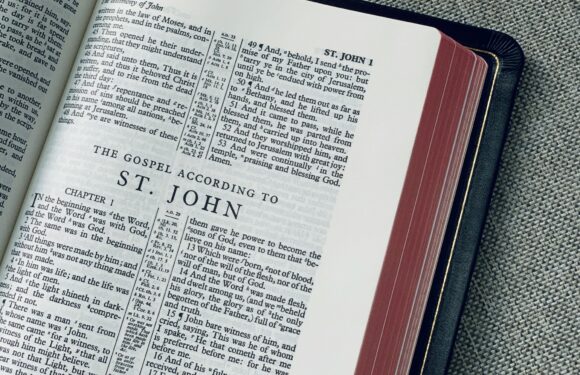 Open Bible showing the beginning of the Gospel of John, with a focus on chapter 1, pages slightly curved, placed on a black cover, on a textured gray surface.