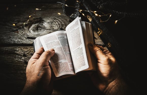 Hands holding an open Bible on a rustic wooden surface, surrounded by string lights and a dark cloth.