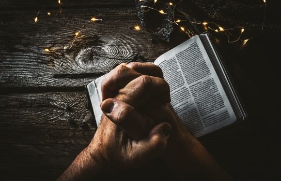 A person holding an open Bible on a dark wooden table, illuminated by a string of small fairy lights, with a knit hat partially visible nearby.