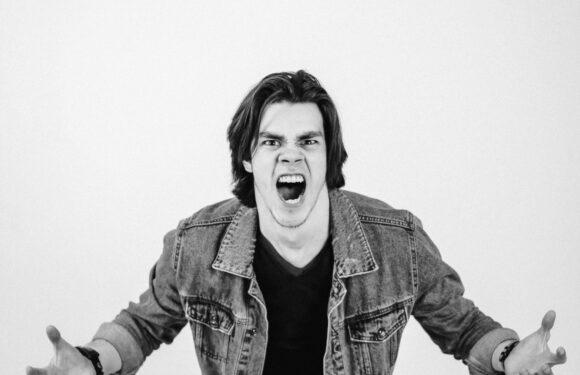 A young man with shoulder-length hair, wearing a denim jacket over a black shirt, appears angry or frustrated, with his mouth open and hands outstretched toward the camera.