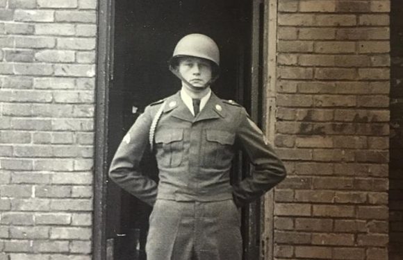 A soldier in uniform with a helmet, standing with hands on hips in front of a brick building entrance with a sign reading "CO B 8 CAV REGT 3RD PLATOON."
