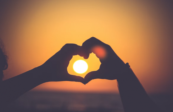 Hands form a heart shape around the setting sun during sunset, with a bright, glowing sky in the background. The image captures a romantic and peaceful moment.