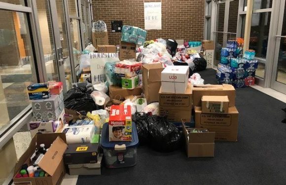 Piles of donation items including boxes of diapers, bottled water, toiletries, and various supplies in a glass-enclosed hallway with a brick wall and a sign that says "Donations Here."