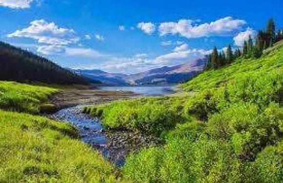 A lush green valley with a small river flowing through, surrounded by hills and dense forests, under a clear blue sky with scattered clouds.