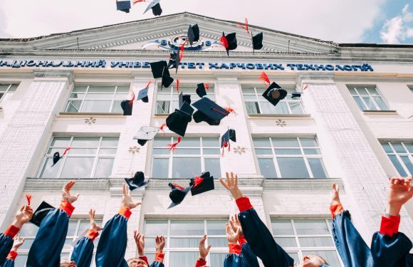 Graduates in blue gowns with red cuffs toss their caps in the air outside a white university building with blue signage.