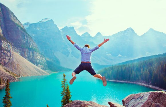 Person jumping with arms and legs spread wide over a mountain lake, surrounded by forest and snow-capped peaks, under a bright blue sky.