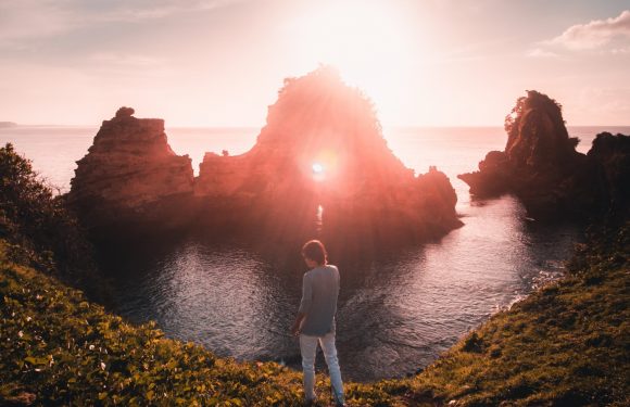 A person stands on grassy cliffs overlooking a rocky coastline at sunset, with the sun shining brightly over the water and creating a warm, glowing atmosphere.