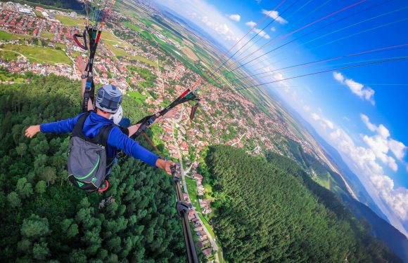 A person paragliding above a green forested area, with a view of a town below, holding onto the control bar and wearing a helmet and blue jacket.