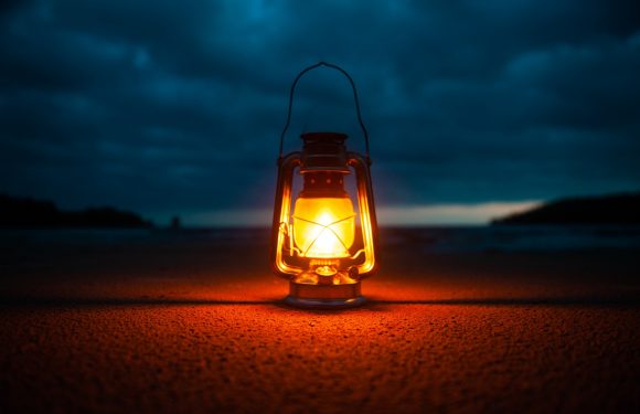 A lantern glowing with warm light sits on the sandy beach during dusk, with dark clouds and a distant shoreline visible in the background.