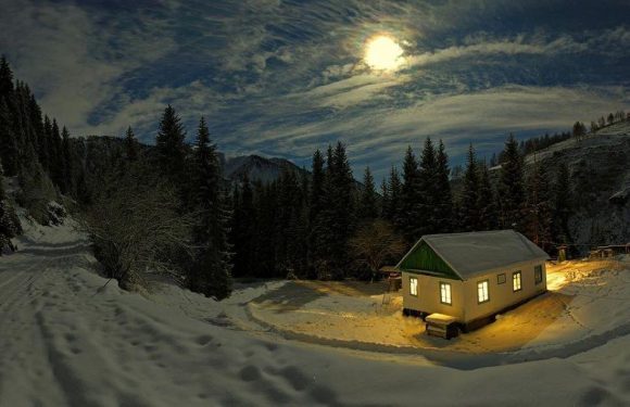 A snow-covered house with lit windows sits beside a wooded mountain landscape under a moonlit sky. The scene is illuminated by the moon and indoor lights.