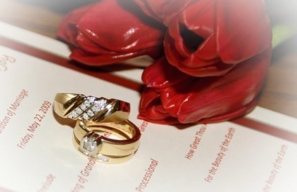 A gold ring with a large central diamond and smaller diamonds on the band rests on a document, alongside a red tulip, on a wooden surface.