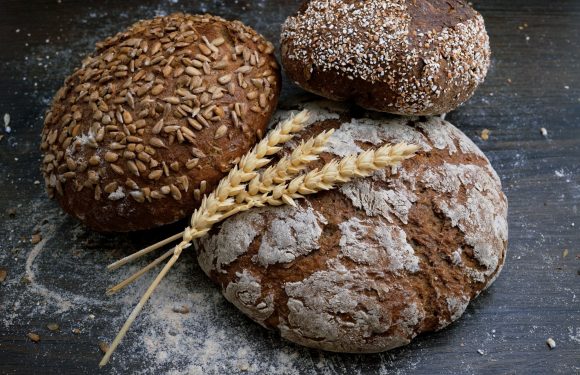 Three loaves of bread with different toppings sit on a dark wooden surface, with two wheat stalks lying on the front loaf.