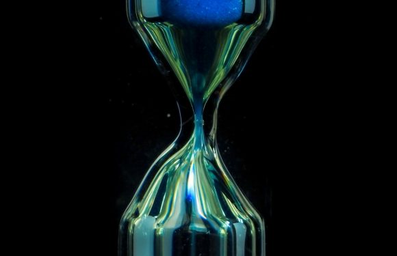 A glass hourglass with blue sand flowing from the top chamber to the bottom against a dark background.
