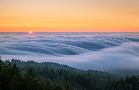 A sunset over a forest with clouds rolling over the hills, creating a surreal, wave-like pattern in the sky above the treetops.