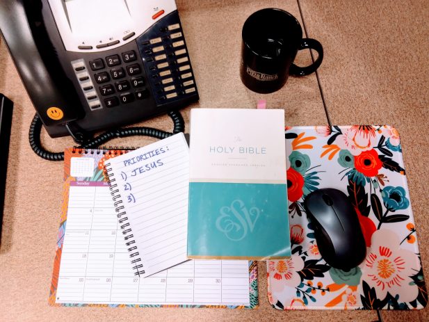 Office desk with phone, coffee mug, computer mouse, calendar, bible, and notebook with the words Priorities and number 1 says Jesus