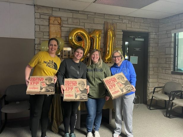 4 ladies holding Pizza Ranch pizza boxes in front of them posing for a group photo in front of gold 9-1-1 shaped balloons.