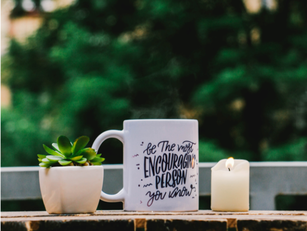 Succulent in a small square pot next to a coffee mug that says "be the most encouraging person you know" next to a burning candle stick all set on a brick in an outdoor setting