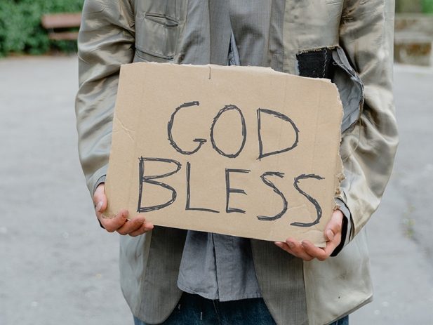 Person outside on a sidewalk holding a handwritten cardboard sign that reads "God Bless".