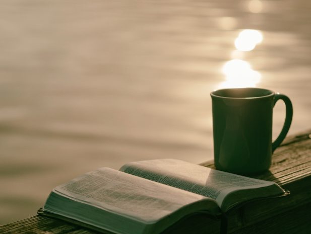 Open Bible and cup of coffee next to lake.
