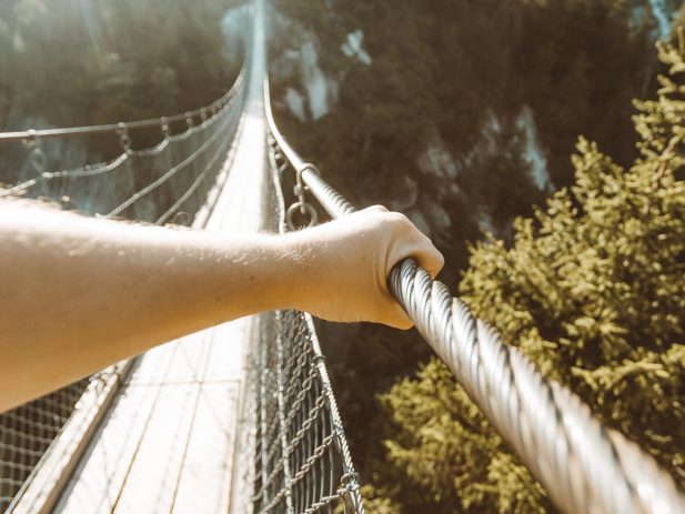 Arm of a person holding onto a rope hand rail of a walking bridge over mountains