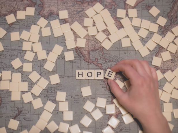 Flat surface with a world map laid out and upside down scrabble tiles spread across the area. in the middle of the tiles is a hand overturning tiles to spell the word hope.
