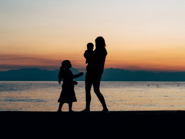 Silhouette of a woman holding infant with young child standing next to them on the beach in front of a sunset.