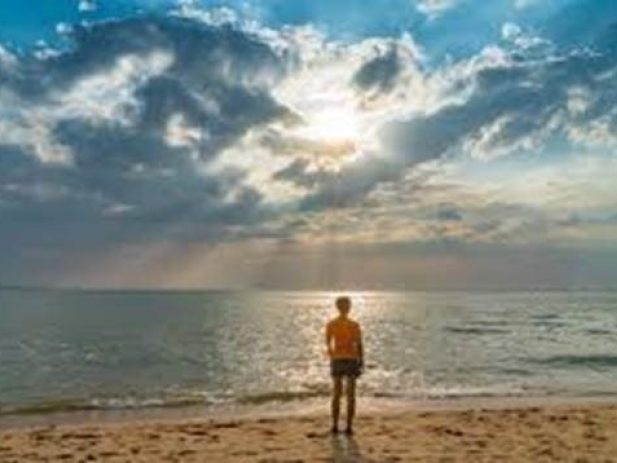 Person standing on the beach in front of the ocean with the sun shining through clouds in the background.