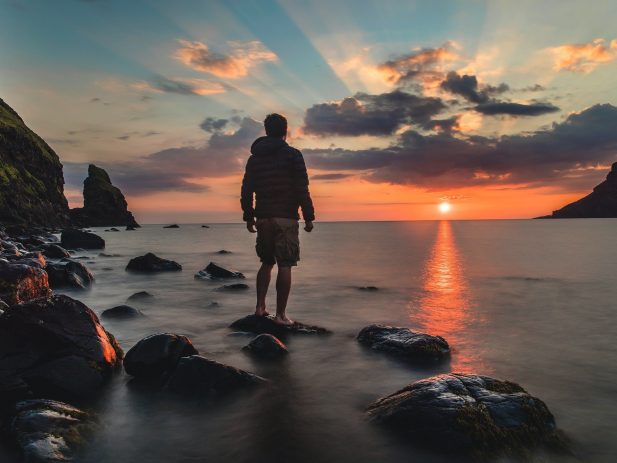 man standing on rocks in the water facing away from the camera looking at a sunset over a large body of water