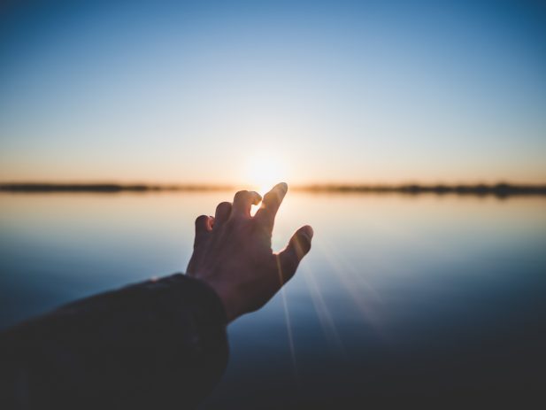Hand reaching out to sun over a lake.