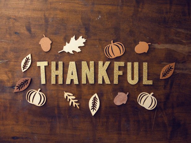 word thankful in gold glitter stickers on a piece of wood with paper shapes of leaves, pumpkins and acorns around it.