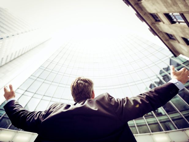 Man in work suit with arms outstretched to the sides looking up to the sky in between skyscrapers