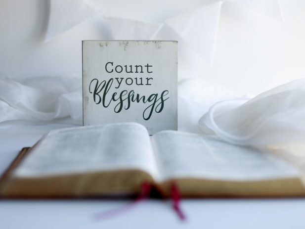 Open bible on a white surface with a white blanket tossed around and a decorative sign that reads count your blessings against a white wall