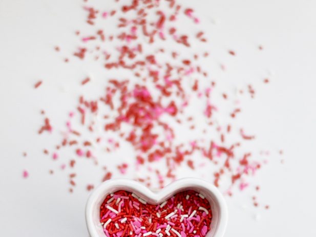 White background with small white heart-shaped bowl filled with red, pink and white sprinkles and some of the sprinkles spilled around the bowl