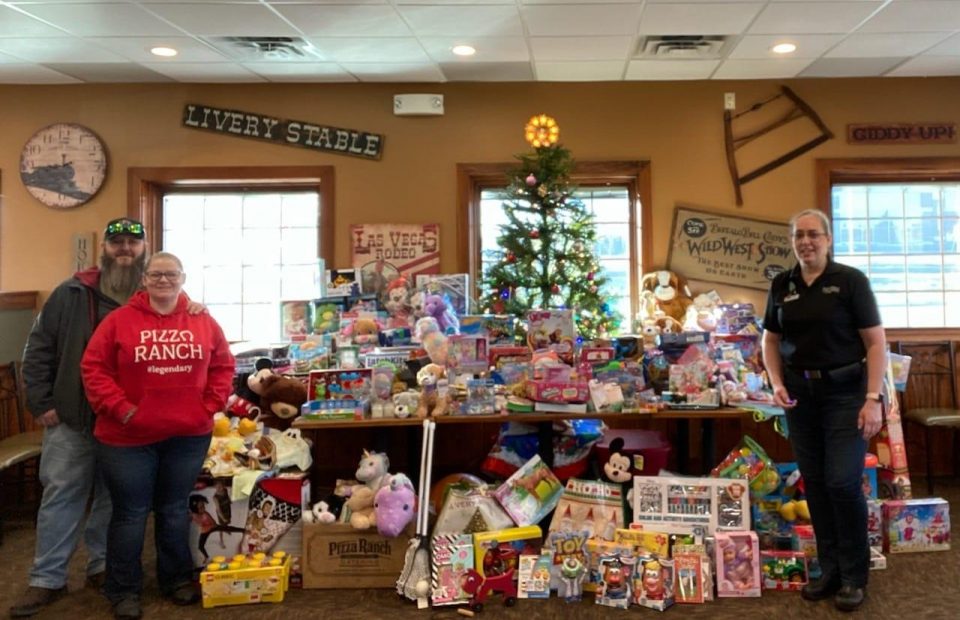 Three people standing behind a large pile of toys and stuffed animals in a room decorated with vintage signage and a Christmas tree.