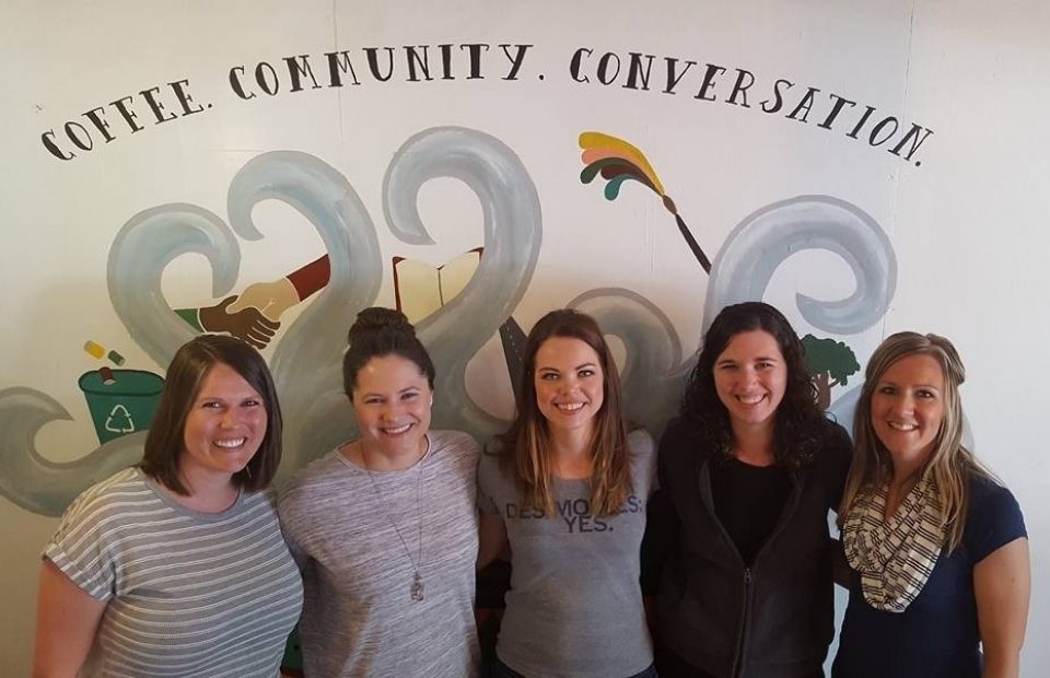 Five women standing together in front of a wall with a mural that includes the words "Coffee. Community. Conversation." and various illustrations.