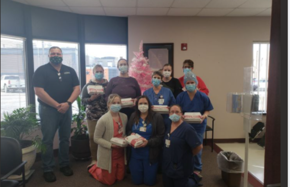 Group of nine people, some in uniforms, wearing masks, holding boxes, standing in a room with a pink Christmas tree, window, and plants.