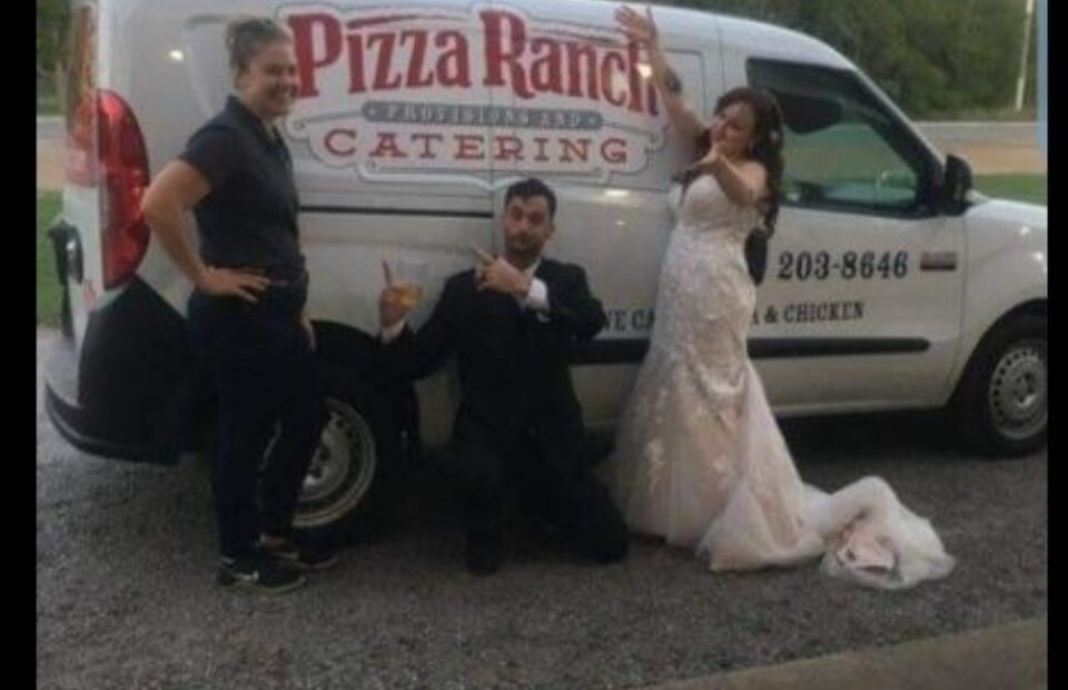 A woman in a wedding dress, a man in a suit, and a worker in a Pizza Ranch uniform pose in front of a catering van with trees and an overcast sky in the background.