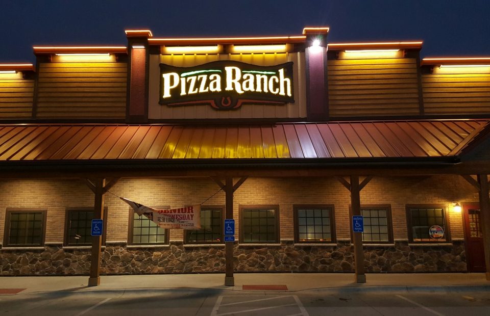 A restaurant building with a large illuminated "Pizza Ranch" sign on the roof, surrounded by a brick and stone facade, with parking spaces in front.