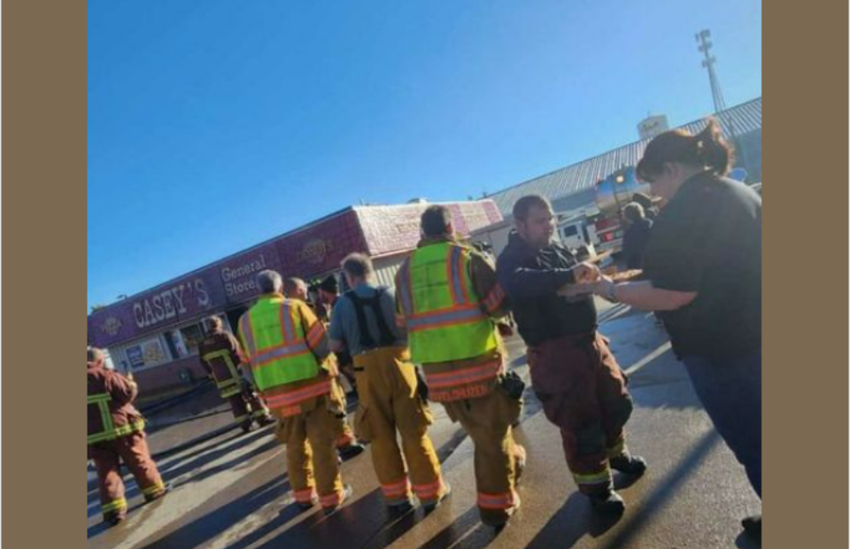 Group of firefighters in reflective gear receive a meal outside a store on a sunny day, with their shadows cast on the ground and a clear blue sky above.