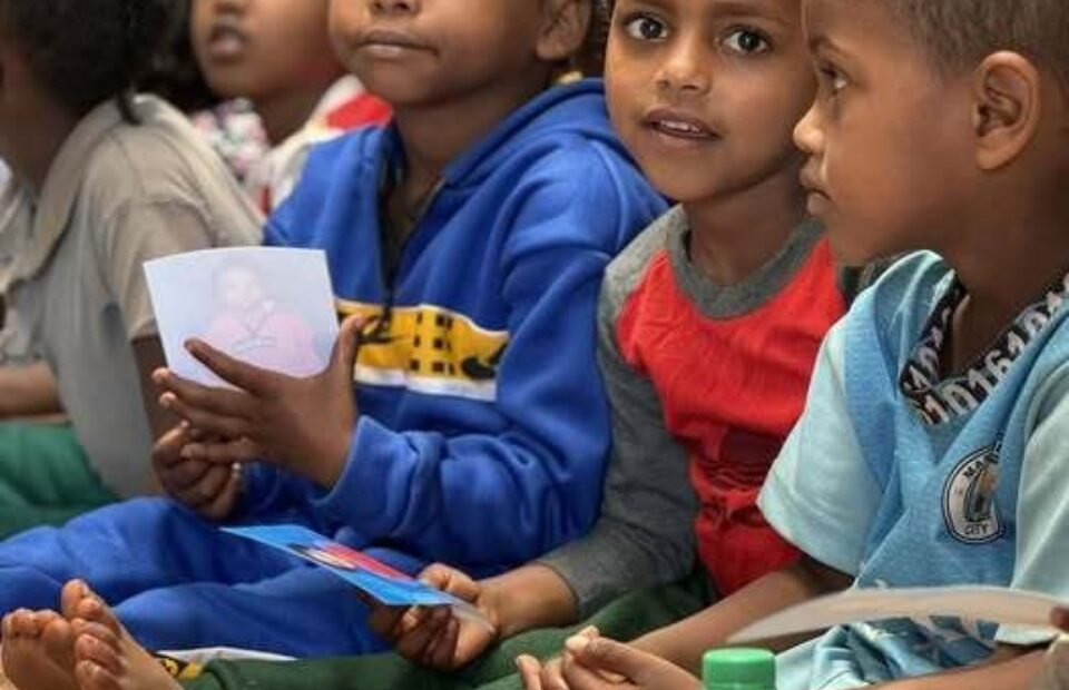 Group of children sitting together indoors on a colorful mat, holding papers and a bottle of beverage, with their legs crossed and attentive expressions.