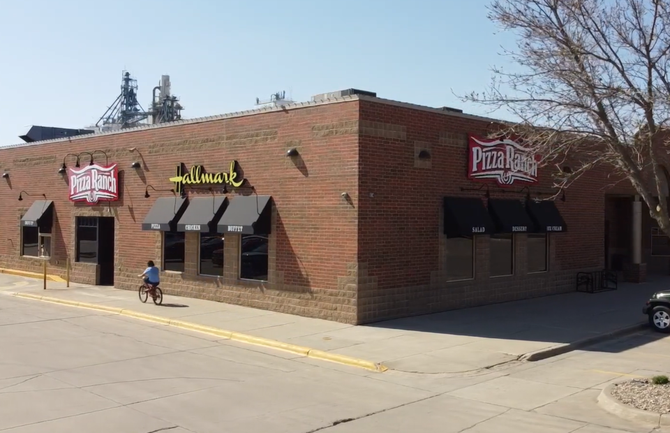 A person riding a bike in front of a large Pizza Ranch restaurant with "Fallmark" signage, brick exterior, and black awnings; leafless tree and parked car nearby.