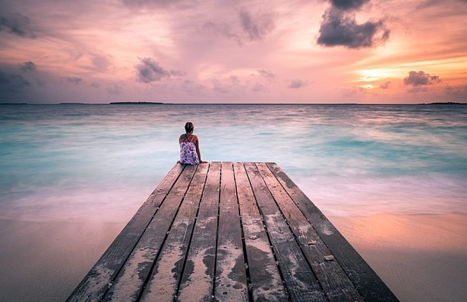 Girl sitting on a wooden dock overlooking the ocean at sunset, with colorful clouds and calm waves. She has a backpack and is facing away from the camera.