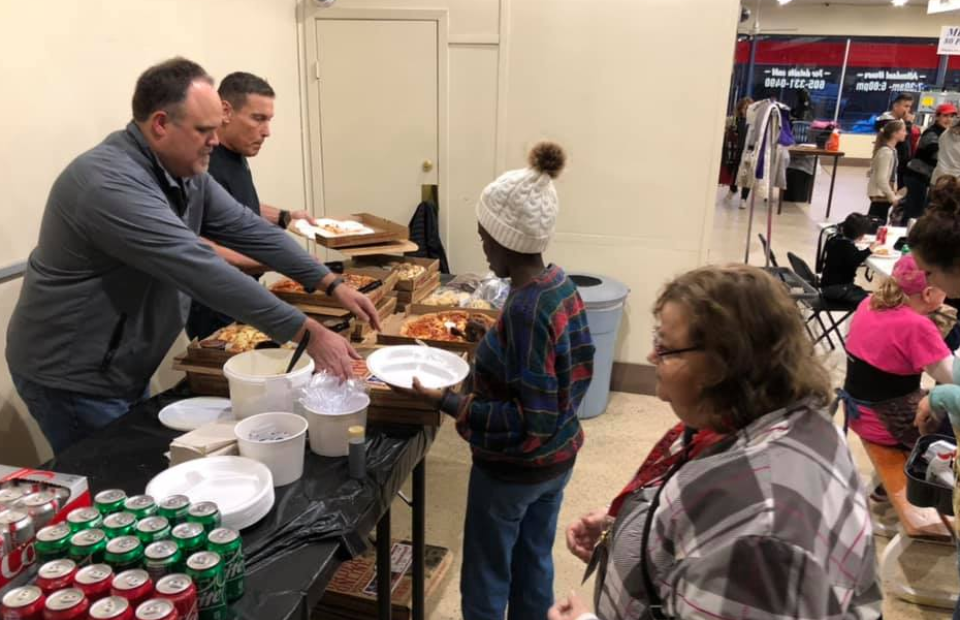 Two men are serving pizza at a buffet-style table, while a young girl in a colorful sweater and knit hat waits with an adult woman nearby. Other people are seated at tables in the background.