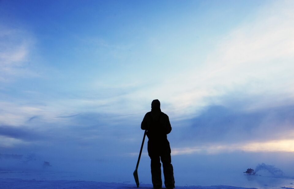 A person stands on snow or ice holding a tool, looking towards a cloudy sky with a boat in the distance on a frozen landscape.