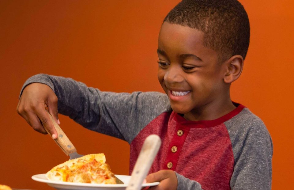 A smiling boy in a red and gray shirt is serving himself a slice of pizza onto a white plate. The background is a warm orange color.