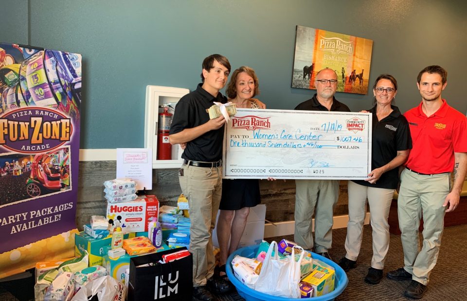 Group of five people holding a large donation check to a Women's Care Center, standing behind a table with diaper supplies and a "FunZone Arcade" sign.